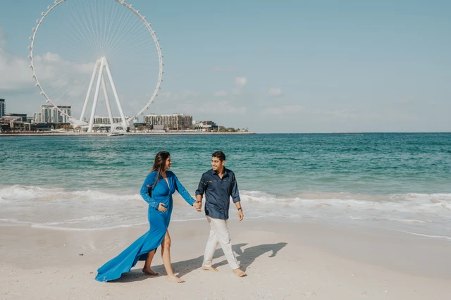 A couple walks hand in hand on Dubai beach with Ain Dubai visible behind them during a beach photoshoot.