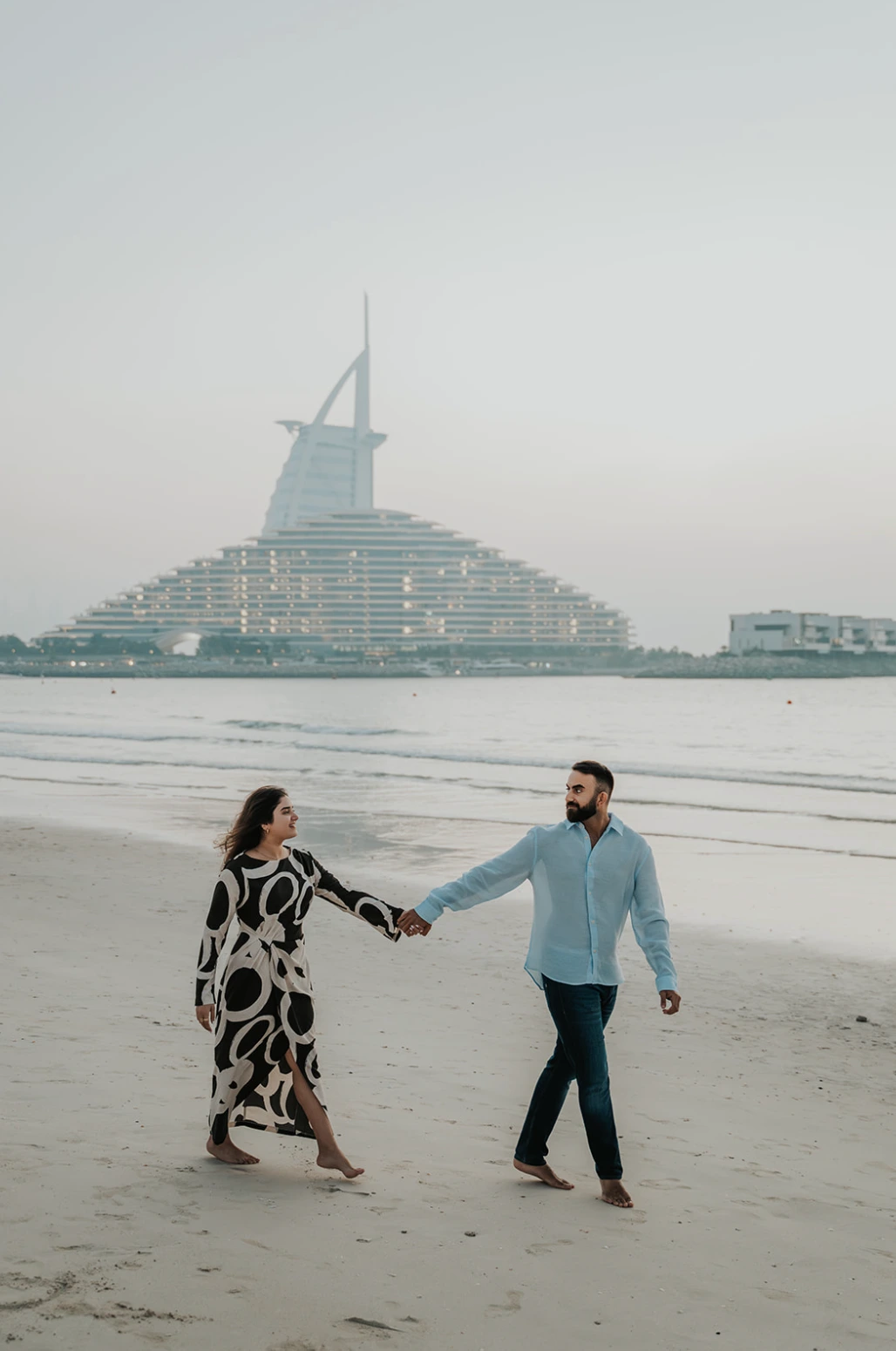 A couple walks near the water with Jumeirah Beach Hotel in the background, illustrating beach timing options in Dubai.