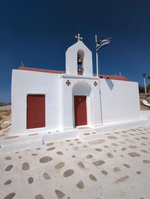 Small white Mykonos chapel with a red roof, twin bells, red doors and a Greek flag