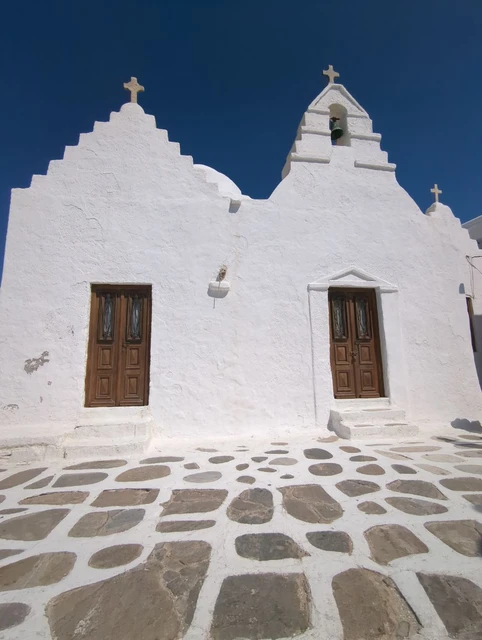 Sculptural white walls and brown doors of Panagia Paraportiani beneath a clear blue sky in Mykonos Town