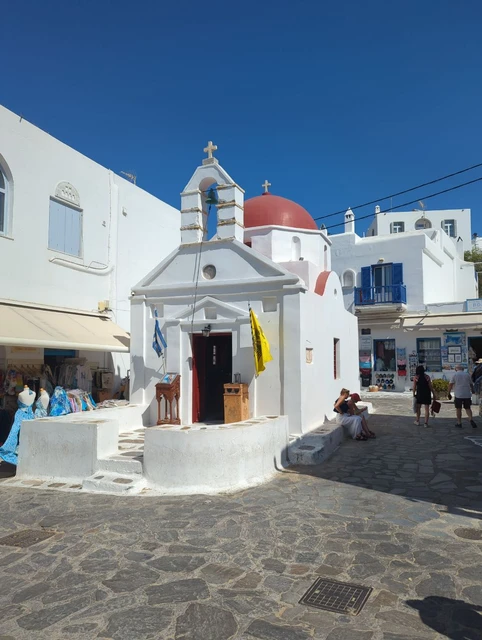 White chapel with a red dome and bell tower rising between shops and pedestrians in Chora