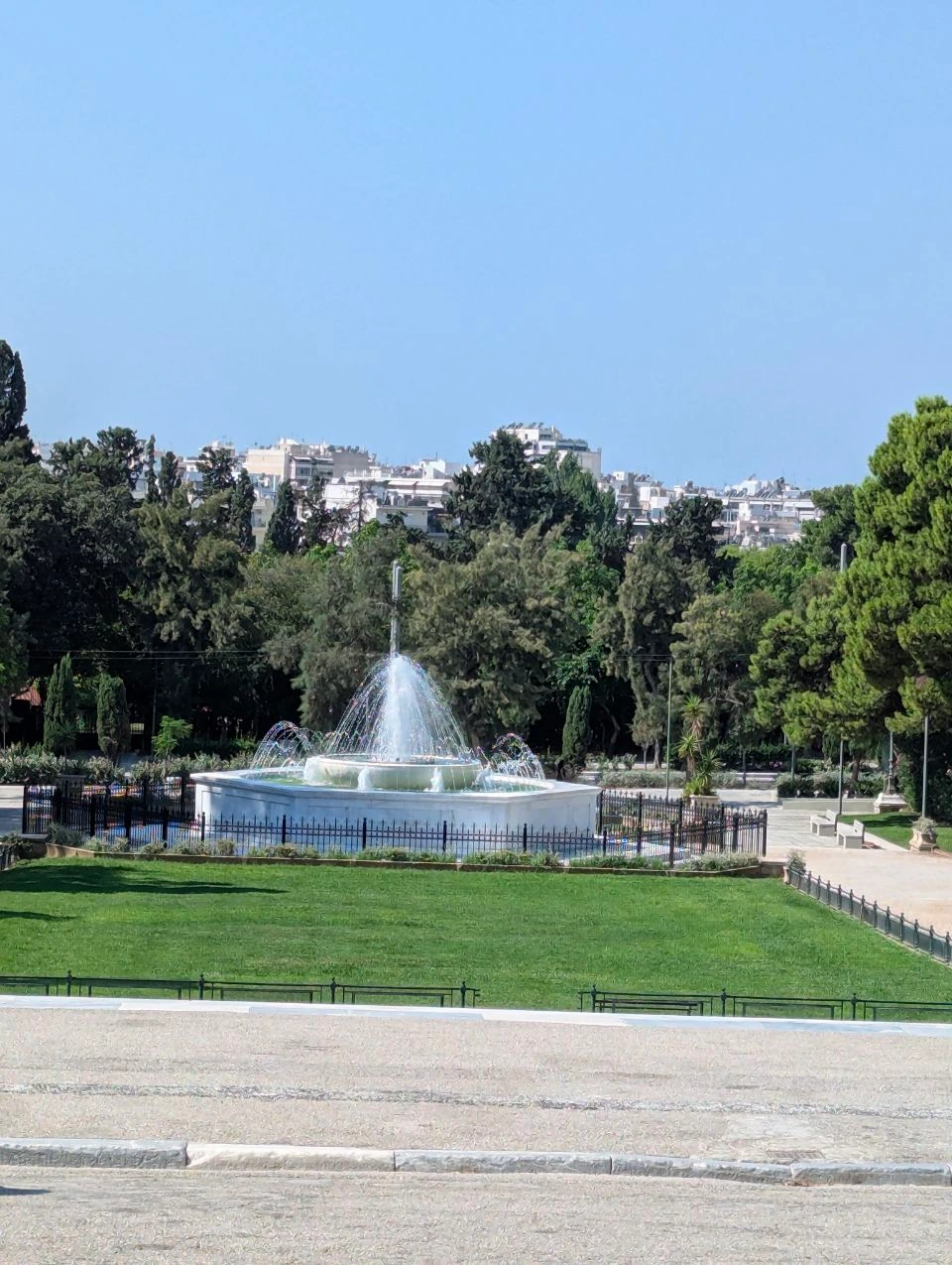 A white fountain sprays above a green lawn surrounded by mature trees near Zappeion in central Athens.