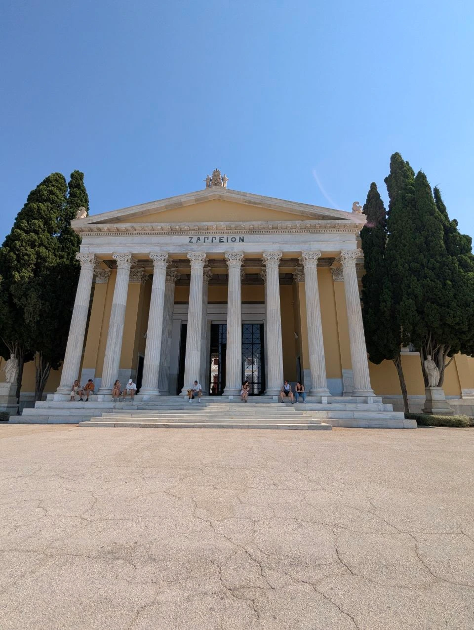 Visitors sit on the broad steps beneath the white columns of Zappeion Hall in Athens.
